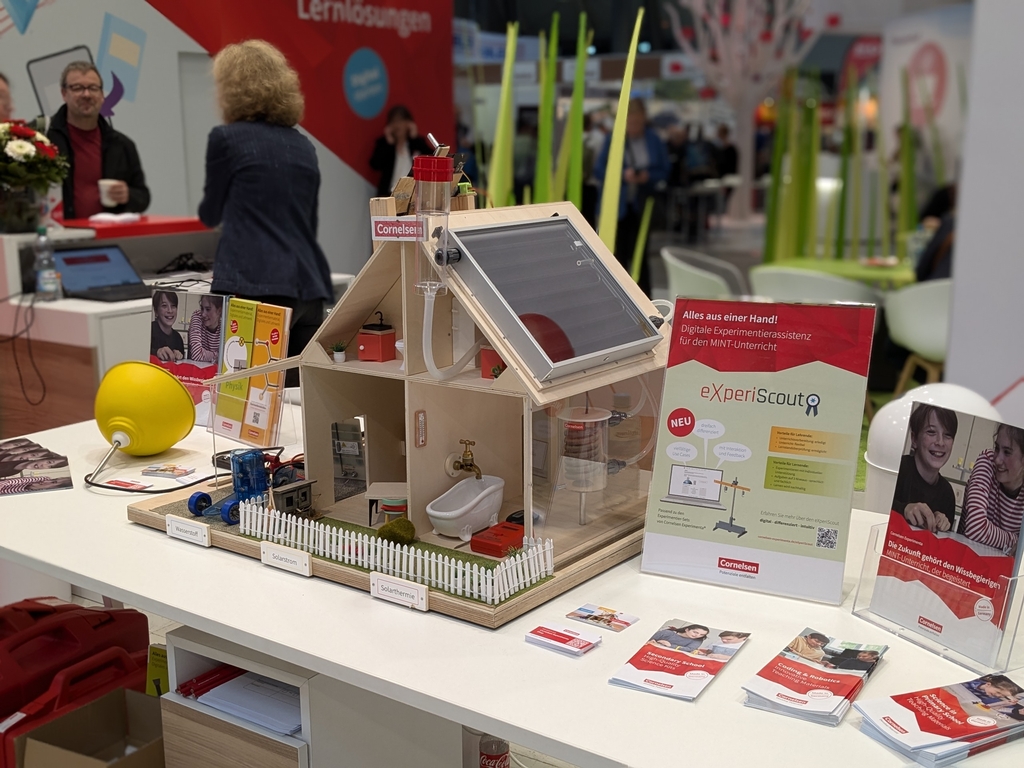 Das Bild zeigt ein Modell eines Energiewandelhauses mit verschiedenen Experimentiermöglichkeiten, das auf einem Messestand für Bildungsbedarf ausgestellt ist. Daneben stehen Informationsmaterialien zur digitalen Unterstützung im MINT-Unterricht.