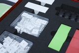 Das Bild zeigt eine transparente Box mit vielen kleinen weißen Bauelementen, die ordentlich auf einem schaumstoffhaltigen Untergrund angeordnet sind. Darüber hinaus sind mehrere farbige Platten und Gummireifen sichtbar, die Teil eines Experimentierkits für den Bildungsbereich sind.
