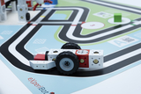 Das Bild zeigt ein Spielzeugfahrzeug, das aus modularen Bausteinen besteht und auf einer Spielmatte mit Fahrstrecke platziert ist. Die Mathe ist farbenfroh gestaltet und weist verschiedene Symbole sowie QR-Codes auf, die für experimentelles Lernen genutzt werden können.