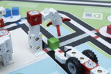 Das Bild zeigt eine Szenen aus einem Lernspielzeug, das aus modularen Bausteinen besteht, die einen Roboterarm und ein Fahrzeug darstellen. Der Roboterarm greift einen grünen Block, während sich die Szenerie auf einem farbenfrohen, spielerischen Spielbrett abspielt.