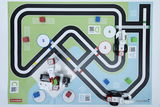 Das Bild zeigt eine Spielmatte mit einer Straßenführung, auf der verschiedene Stationen und farbige Blöcke platziert sind. Zwei Roboterfahrzeuge aus Bauklötzen sind auf der Matte positioniert und bereit, die Strecke zu erkunden.