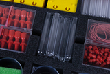 Das Bild zeigt eine Auswahl von Experimentiermaterialien, darunter farbige Kunststoffteile, Glasröhren sowie Gummibänder, die in einem gepolsterten Verpackungsmaterial angeordnet sind. Diese Utensilien sind für den Einsatz im Bildungsbereich konzipiert, um Experimente durchzuführen.