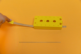 Das Bild zeigt ein gelbes Experimentiergerät mit einer flexiblen Metalllasche und einer geraden Metallstange, die auf einem orangefarbenen Hintergrund liegen. Das Gerät scheint für Bildungszwecke konzipiert zu sein und könnte für physikalische Experimente verwendet werden.