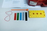 Das Bild zeigt Materialien für Experimente zu Klängen und Geräuschen, darunter eine gelbe Platte mit Löchern, farbige Streifen und Gummiringe. Außerdem liegt eine Anleitung auf dem Tisch, die die Durchführung von Versuchen beschreibt.
