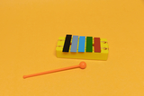 Das Bild zeigt ein buntes Xylophon auf einer gelben Unterlage. Neben dem Xylophon liegt ein orangefarbener Schlägel, der zum Spielen des Instruments verwendet wird.