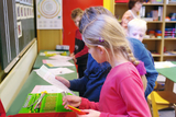 Das Bild zeigt eine Gruppe von Kindern in einem Klassenzimmer, die eine Experimentierstation nutzen. Zwei Kinder betrachten gemeinsam eine Anleitung, während im Hintergrund weitere Schüler und ein Lehrer zu sehen sind, die an einem Tisch arbeiten.