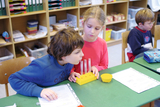 Das Bild zeigt zwei Kinder, die an einem Tisch mit Experimentiermaterial arbeiten. Sie konzentrieren sich auf ein gelbes Reagenzglas-Set und scheinen an einem naturwissenschaftlichen Experiment interessiert zu sein.