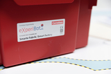 Das Bild zeigt einen roten Karton mit der Bezeichnung "eXperiBot" und dem Zusatz "Komplett-Set/Smarte Fabrik". Er befindet sich auf einem Untergrund mit einer blauen Fläche und einer gestrichelten Linie, die wahrscheinlich Teil eines Experimentier- oder Lernsets ist.