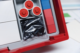 Das Bild zeigt einen Organizer mit verschiedenen Experimentiermaterialien, darunter drei runde Behälter mit roten Köpfen und eine Tüte mit schwarzen Bauteilen. Neben diesen befinden sich auch farbige Kunststoffsteine in Blau und Rot.