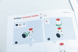 Das Bild zeigt eine Anleitung zum Aufbau eines "Smarter Greifer", in dem verschiedene Bauteile wie Räder und Würfel dargestellt sind. Schritt-für-Schritt-Illustrationen verdeutlichen, wie die Komponenten miteinander verbunden werden.