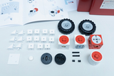 Das Bild zeigt verschiedene Bauteile eines Experimentier-Sets, darunter Würfel mit verschiedenen Funktionen, Räder und Konstruktionselemente. Dazu liegt ein Anleitungsheft, das die Montage und experimentelle Nutzung der Teile beschreibt.