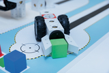 Das Bild zeigt ein technisches Modell, das aus einem Fahrzeug mit großen Gummireifen und einer grünen Würfelkomponente besteht. Es steht auf einem farbigen Untergrund mit verschiedenen geometrischen Formen und Linien, die offenbar einem Lern- oder Experimentierszenario dienen.