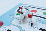 Das Bild zeigt ein zusammengestelltes Roboter-Modell aus Bauklötzen, das auf einer bedruckten Unterlage platziert ist. Die Unterlage enthält verschiedene Markierungen und Bereiche, die vermutlich für Programmier- oder Trauingszwecke genutzt werden.
