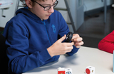 Ein junger Schüler sitzt an einem Tisch und hält ein schwarzes Rad in der Hand. Auf dem Tisch liegen zudem mehrere farbige, kubusartige Bauteile, die wahrscheinlich für experimentelle Zwecke im Bildungsbereich verwendet werden.