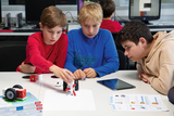 In dem Bild arbeiten drei Kinder konzentriert an einem Tisch, während sie an einem kleinen, selbstgebauten Roboter basteln. Auf dem Tisch sind auch einige Bücher und Tablets zu sehen, die das Experimentieren unterstützen.