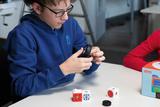 Das Bild zeigt einen Jungen, der konzentriert an einem Experimentierbausatz arbeitet. Um ihn herum sind verschiedene Bauteile platziert, während im Hintergrund eine weitere Person und eine Verpackung zu sehen sind.