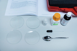 Vier Glasschalen liegen übersichtlich auf einem Tisch, umgeben von verschiedenen Chemikalien und Laborausrüstung, darunter ein Löffel, eine Pipette und ein Behälter mit Zucker. Im Hintergrund ist eine Bedienungsanleitung sichtbar, die Anweisungen für Experimente gibt.