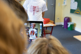 Ein Schüler steht vor einer Kamera und trägt ein T-Shirt, das ein anatomisches Herz zeigt. Im Hintergrund ist ein Klassenraum mit Tischen, Stühlen und Büchern zu sehen, während ein anderer Schüler das Geschehen über ein Tablet aufnimmt.