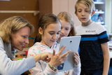 Eine Gruppe von Kindern und einer Erwachsenen beobachtet aufmerksam ein Tablet, auf dem etwas Spannendes angezeigt wird. Die Szene findet in einem Klassenzimmer statt und vermittelt eine freundliche Lernatmosphäre.