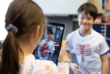 Ein Kind hält ein Tablet, auf dessen Bildschirm ein zweites Kind mit einem T-Shirt zu sehen ist, das anatomische Merkmale zeigt. Die Szene spielt in einem Klassenzimmer und dient vermutlich einem experimentellen Lernzweck.