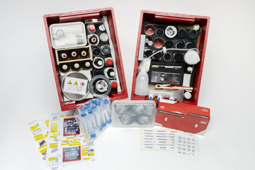 Zwei rote Kisten enthalten eine Vielzahl von Experimentiermaterialien, einschließlich Testschläuchen, chemischen Reagenzien und einem Sicherheitslabel. Zusätzlich sind Anleitungen und Informationsblätter für verschiedene Experimente auf einem Tisch verteilt.