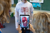 Ein Kind steht vor einer Tafel, trägt ein T-Shirt mit anatomischem Druck und wird durch ein Tablet betrachtet, das eine digitale Darstellung der inneren Organe zeigt. Eine Person hält das Tablet und interagiert mit der Augmented-Reality-Anwendung, um das Modell zu präsentieren.