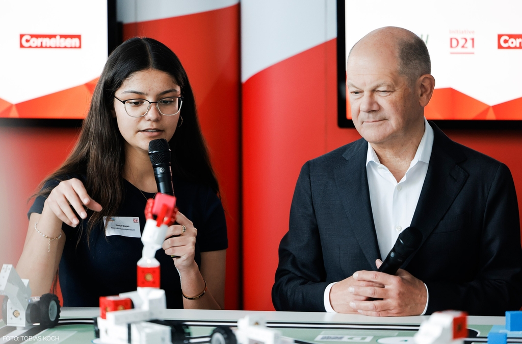 Eine junge Frau präsentiert den Lernroboter eXperiBot mit einem Mikrofon in der Hand, während Bundeskanzler Scholz aufmerksam zuschaut. Im Hintergrund sind Logos und eine kreative Umgebung sichtbar, die auf eine Bildungs- und Innovationsveranstaltung hinweisen.