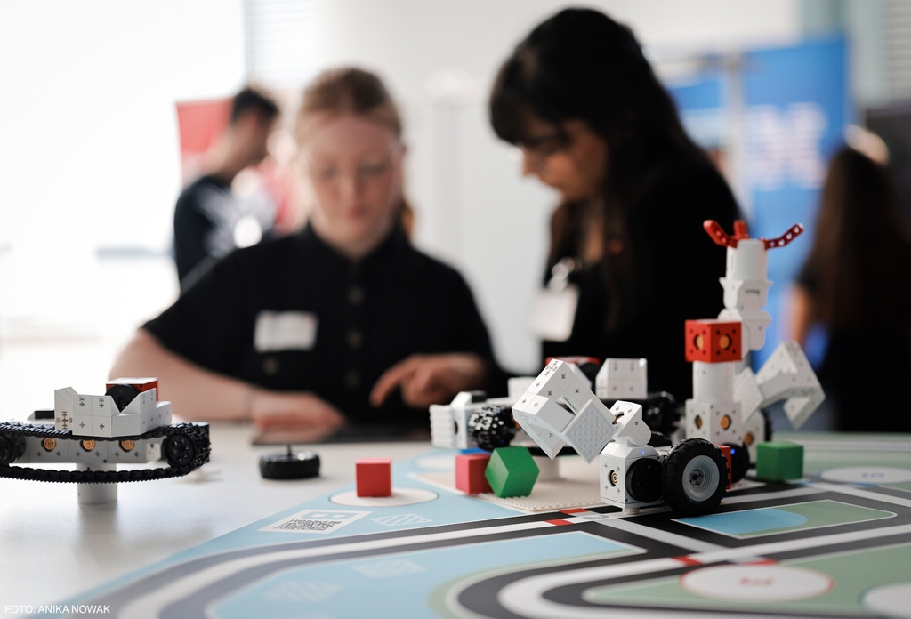 Auf dem Tisch sind verschiedene Lernroboter auf einer Matte platziert., Im Hintergrund sind junge Frauen zu sehen, die sich mit Programmieren der Lernrobotern beschäftigen und an einem Lernprojekt arbeiten.
