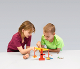 Zwei Kinder interagieren mit einem Spielzeugwaage, wobei sie aufmerksam beobachten, wie verschiedene Objekte ihre Balance beeinflussen. Rund um die Waage liegen unterschiedliche Materialien, die sie verwenden, um Experimente durchzuführen und zu lernen.