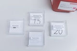 Vier transparente Kästchen sind auf einem Tisch angeordnet, die jeweils mit Bezeichnungen wie "Paare finden" und Zahlenkarten für Hundertkette und Zahlenstrahl versehen sind. Die Kästchen enthalten Materialien zur Förderung mathematischer Fähigkeiten im Bildungsbereich.