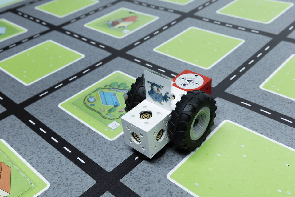 Ein fahrzeugähnliches Modell mit großen Reifen steht auf einem matten Untergrund, der Straßen und grüne Bereiche darstellt. Das Modell zeigt bunte Würfel mit Symbolen und einem Bild eines Vogels auf der Rückseite.