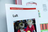 Eine Seite eines Lehrbuchs für Experimente zeigt das Thema „Luft“ und illustriert es mit einem Bild von zwei Kindern, die mit bunten Bällen spielen. Im Hintergrund sind weitere Kinder und Materialien zu sehen, die eine experimentelle Lernumgebung schaffen.