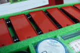 Im Bild sind mehrere rote Bauteile mit Rädern zu sehen, die in einem schaumstoffgefüllten Kasten angeordnet sind. Daneben liegen verschiedene Utensilien wie Vaseline und Rohre, die für experimentelle Zwecke verwendet werden können.