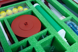 In einem gut organisierten Kasten befinden sich verschiedene Experimentiermaterialien, darunter gelbe Kugeln, rote Scheiben und ein grüner Stab. Die Fächer sind mit einer schaumstoffartigen Polsterung ausgekleidet, die die Materialien schützt und ordentlich hält.