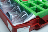 Ein roter Kasten enthält verschiedene Experimentiervorbereitungen, darunter mehrere transparente Trichter und Gefäße, die in einer schützenden grünen Schaumstoffeinlage angeordnet sind. Zusätzlich sind einige Materialien zum Experimentieren, wie beispielsweise Papier, im Inneren sichtbar.