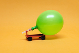 Auf einem orangefarbenen Hintergrund steht ein kleines, rotes Modellfahrzeug, an dem ein grüner Ballon befestigt ist. Der Ballon wird durch einen Schlauch mit dem Fahrzeug verbunden, was auf ein Experiment zur Demonstration von Luftdruck oder Antriebskraft hinweist.
