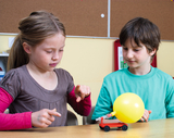 Zwei Kinder experimentieren zusammen mit einem kleinen roten Wagen, der mit einem aufgeblasenen gelben Ballon verbunden ist. Sie beobachten gespannt, wie der Wagen sich bewegt, während sie neugierig auf das Experiment eingehen.