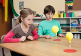 Zwei Kinder sitzen an einem Tisch und beobachten zwei gelbe Ballons, die auf roten Ständern stehen. Sie scheinen fasziniert von einem Experiment zu sein, das offensichtlich auf einfache physikalische Prinzipien abzielt.