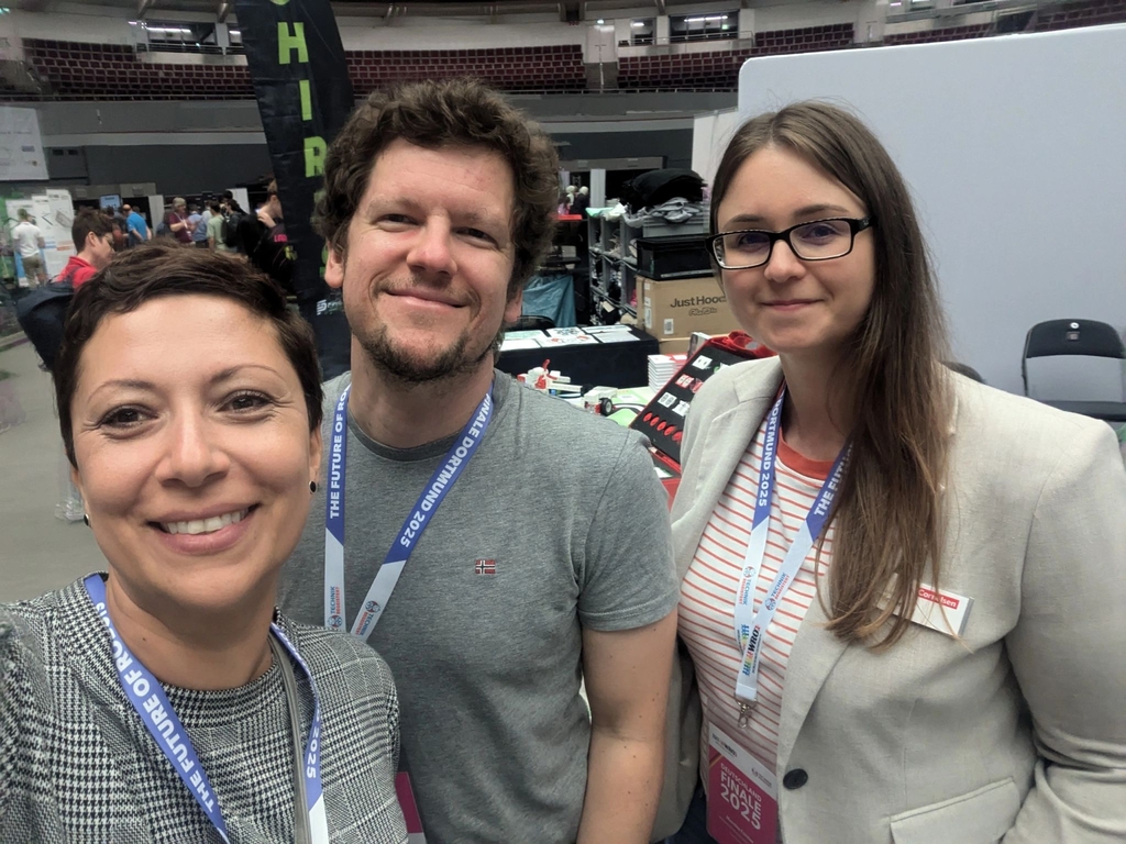 Drei Personen lächeln in die Kamera und scheinen an einem Stand bei einer Veranstaltung für Experimentierbedarf beteiligt zu sein. Im Hintergrund sind Stände und verschiedene Produkte zu erkennen, die für Bildungszwecke angeboten werden.