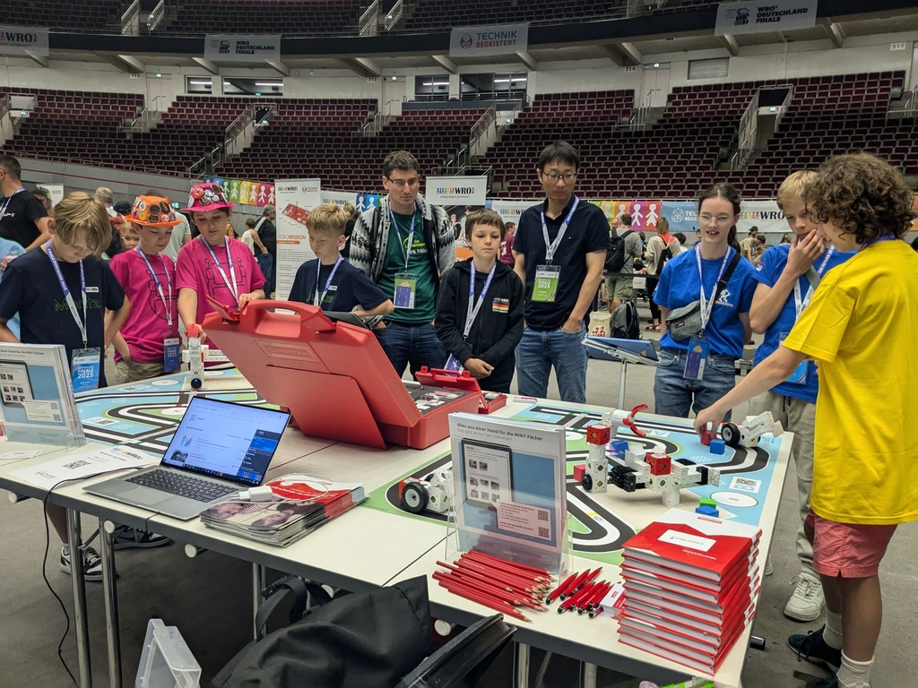 Eine Gruppe von Kindern und Jugendlichen steht um den Cornelsen Experimenta Tisch, der mit dem eXperiBot Smart Team und zwei Spielmatten ausgestattet ist. Sie sind interessiert am eXperiBot Lernroboter, der auf einer farbig gestalteten Matte fährt.