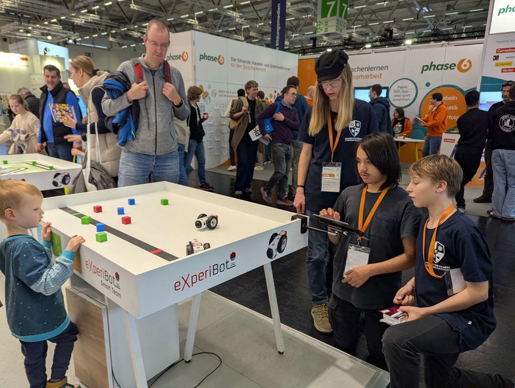 Auf einem Tisch werden kleine Roboter pr&auml;sentiert, die auf einer festgelegten Bahn spielen. Interessierte Zuschauer, darunter ein kleines Kind und mehrere Erwachsene, beobachten gespannt die Demonstration des Roboters und dessen Interaktionen mit farbigen Formen.