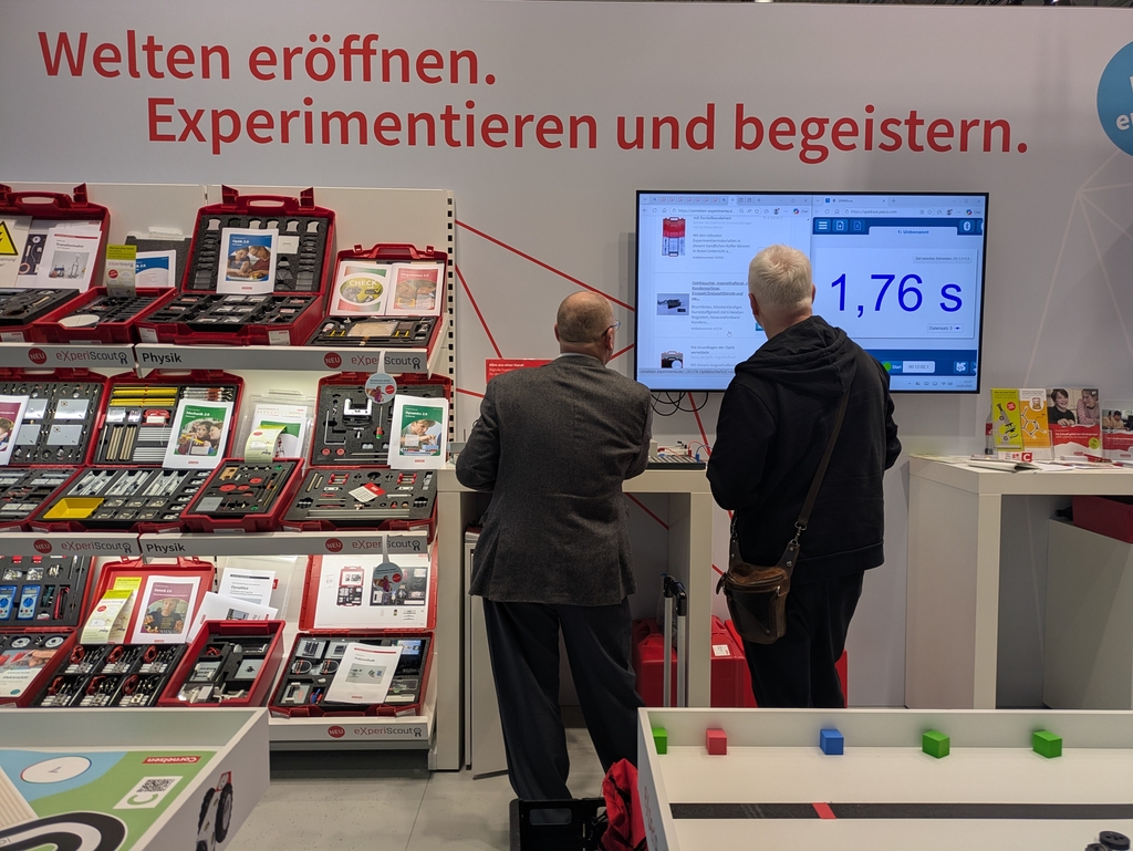 Zwei Personen stehen vor einem gro&szlig;en Bildschirm, auf dem eine Zeitmessung von 1,76 Sekunden angezeigt wird. Im Hintergrund sind Regale mit Experimentierk&auml;sten und Materialien f&uuml;r den Physikunterricht sichtbar, die zum Experimentieren einladen.