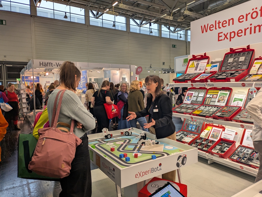 Eine Pr&auml;sentatorin erkl&auml;rt den Besuchern den eXperiBot, ein interaktives Lernspielzeug, das f&uuml;r den Einsatz im Bildungswesen konzipiert ist. Im Hintergrund sind verschiedene Experimentierkits sowie Materialien f&uuml;r den naturwissenschaftlichen Unterricht ausgestellt.