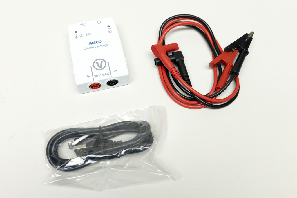 Ein kabelloses Spannungsmessgerät von PASCO ist abgebildet, das für Experimente im Bildungsbereich verwendet wird. Neben dem Messgerät liegen rote und schwarze Krokodilklemmen sowie ein USB-Kabel in einer Verpackung.