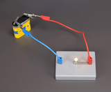 An einem Batteriehalter sind zwei Kabel angeschlossen, eines rot und eines blau. Diese Kabel führen zu einer kleinen Glühbirne auf einer grauen Plattform, die leuchtet, wenn die Schaltung geschlossen ist.