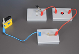 Ein Batterieanschluss mit Kabeln ist zu sehen, der mit mehreren grauen Experimentierplatten verbunden ist. Diese Platten enthalten verschiedene elektrische Komponenten, darunter eine Lampe und einen Druckknopf, die für experimentelle Zwecke im Bildungsbereich verwendet werden.