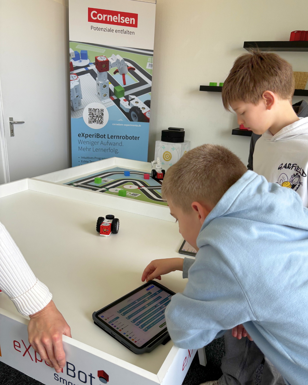 Zwei Kinder interagieren mit einem programmierbaren Lernroboter auf einem Tisch, während sie ein Tablet nutzen, um Befehle einzugeben. Im Hintergrund ist ein Banner zu sehen, das auf die Lernmöglichkeiten des eXperiBot hinweist.