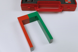 Ein hufeisenförmiger Magnet in Rot und Grün liegt auf einer weißen Fläche. Im Hintergrund ist ein Teil einer roten Kunststoffbox zu sehen.