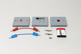 Dargestellt ist ein Set aus drei grauen Modulen mit verschiedenen Schaltern und Kontakten. Ergänzt wird das Experimentierkit durch zwei Verbindungskabel, Krokodilklemmen und eine 9V-Blockbatterie.
