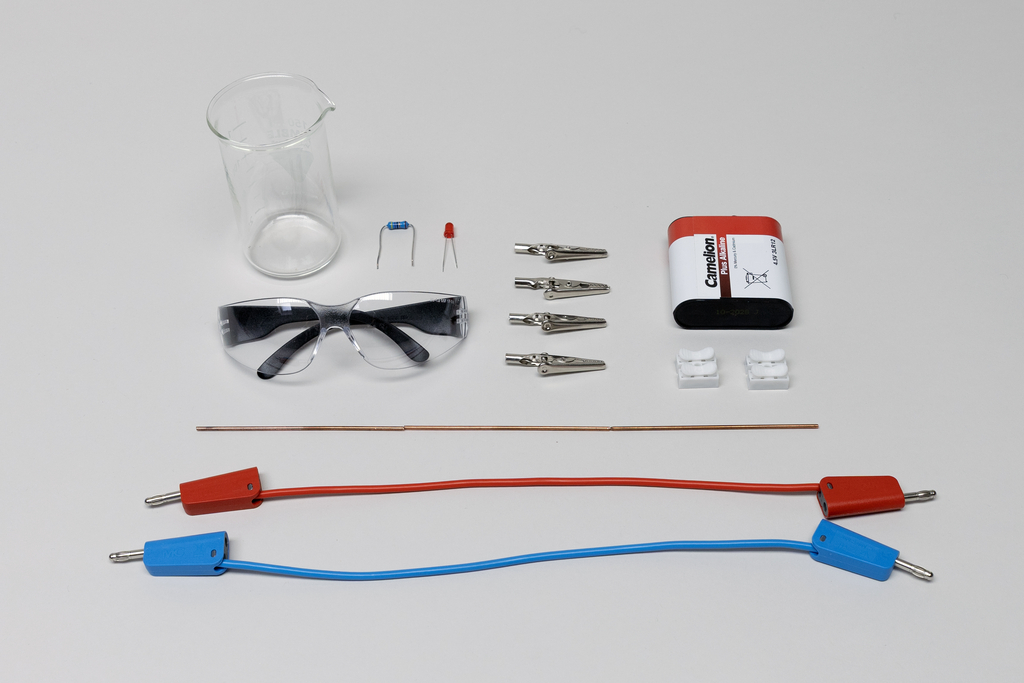 Ein Set aus Laborzubehör umfasst ein Becherglas, eine Schutzbrille, Krokodilklemmen und zwei Anschlusskabel in Rot und Blau. Ergänzt wird die Ausstattung durch eine Batterie, Widerstände und einen dünnen Metallstab auf grauem Hintergrund.